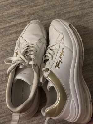 Vita sneakers från Tommy Hilfiger - Snygga vita sneakers från Tommy Hilfiger med gulddetaljer och logga på sidan. Skorna har tjock sula, klassisk snörning och är tillverkade i läder och syntet. Perfekta för dig som vill ha en clean och trendig look.