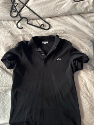 Svart pikétröja från Lacoste - Klassisk svart pikétröja från Lacoste med korta ärmar och den ikoniska gröna krokodilloggan på bröstet. Tröjan har krage och knappar framtill, perfekt för en clean och stilren look.