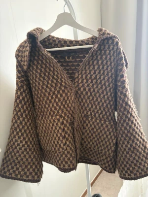 Brun och beige stickad kofta/kappa - Stickad kofta i brunt och beige med rutmönster och två stora knappar framtill. Koftan ånga ärmar och två fickor. Perfekt för dig som gillar elegant stil och vill ha något mysigt och unikt i garderoben.