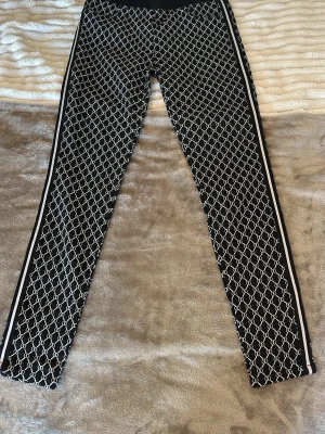 Svarta leggings med kedjemönster - Snygga svarta leggings med vitt kedjemönster över hela byxan och vita ränder längs sidorna. Stretchigt material som sitter tight och bekvämt. Perfekta för dig som vill ha en trendig och sportig look.