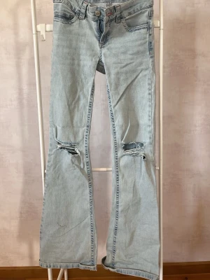 Ljusblå Nelly bootcut jeans med slitningar - Säljer ett par ljusblå bootcut jeans med slitna hål på båda knäna och dekorativa sömmar på bakfickorna. Jeansen har klassisk femficksdesign och är tillverkade i denim. Perfekta för en avslappnad och trendig look.