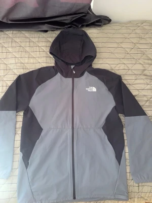 Grå vindjacka från The North Face - Snygg tvåfärgad vindjacka från The North Face i mörkgrått och ljusgrått. Jackan har huva, hel dragkedja framtill och två sidofickor. Tillverkad i lätt och vindtätt material, perfekt för blåsiga dagar. Logotypen syns tydligt på bröstet.