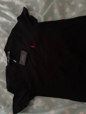 Svart t-shirt från Polo Ralph Lauren - Svart t-shirt från Polo Ralph Lauren med klassisk passform och korta ärmar. T-shirten har en liten röd broderad logotyp på bröstet och är tillverkad i mjuk bomull. Perfekt för en stilren och enkel look.