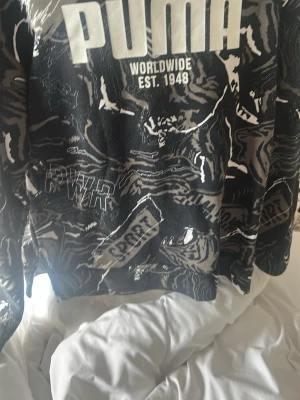 Svart hoodie från Puma med tryck - Cool svart hoodie från Puma med stort vitt logotryck på bröstet och grafiskt mönster i grått och vitt. Text som 'PWR' och 'SPORT' finns på framsidan. Hoodien har en avslappnad passform och är tillverkad i mjukt material. Storlek 152