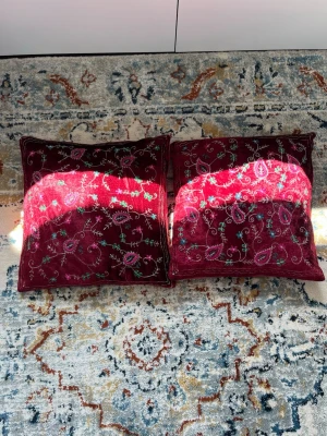 Två vinröda kuddfodral med brodyr - Säljer två fyrkantiga kuddfodral i vinrött sammetstyg med broderade blommönster i rosa, grönt och blått. Kuddfodralen har dekorativa detaljer och ger en färgklick till soffan eller sängen. Perfekt för dig som gillar bohemisk eller orientalisk inredning.
