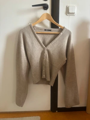 Tröja från Gina Tricot  - Trendig beige cropped kofta med v-ringning och knappar framtill. Koftan har långa, vida ärmar och ribbstickad nederkant som ger en snygg siluett. Perfekt att slänga över en topp för en avslappnad look.