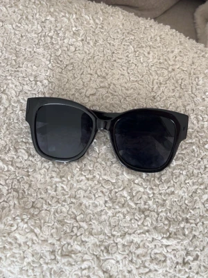 Svarta solglasögon från Gina Tricot - Snygga svarta solglasögon från Gina Tricot med breda bågar och en stilren design. Perfekta för att lyfta vilken outfit som helst och ger en cool vibe. Tillverkade i syntetmaterial.