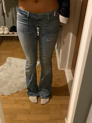 Ljusblå bootcut jeans med slitningar - Säljer ett par ljusblå bootcut jeans med slitningar på låret och knät. Jeansen har stora framfickor och klassiska bakfickor. Materialet är denim och passformen är figurnära upptill med utsvängda ben. Perfekta för en avslappnad och trendig look.