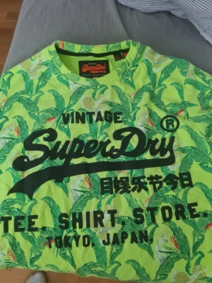 Grön mönstrad Superdry t-shirt - Säljer en grön t-shirt från Superdry med tropiskt bladmönster och stor svart logga framtill. T-shirten har rund hals och korta ärmar. Perfekt för dig som gillar färgstarka plagg och streetwear vibes.