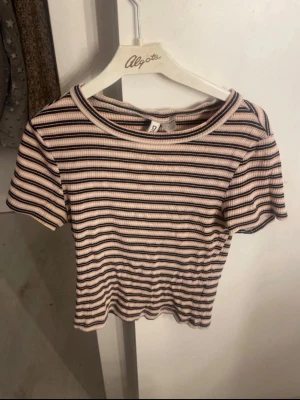 Randig ribbad topp från H&M - Kortärmad topp från H&M i ribbad bomull med smal passform. Mönstret är randigt i beige, svart och rosa toner. Perfekt till jeans eller kjol för en söt outfit