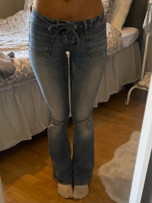 Blå bootcut jeans med snörning - Snygga blå bootcut jeans med cool snörning i midjan och slitningar vid knäna. Jeansen har klassiska fickor fram och bak samt en lätt tvättad look. Perfekta för dig som gillar en trendig och avslappnad stil.