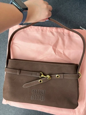 Brun axelremsväska från Miu Miu - Snygg brun handväska från Miu Miu i mjukt läder med präglad logga framtill. Väskan har en justerbar axelrem med guldfärgade metalldetaljer och öljettdekor. Perfekt storlek för det viktigaste och ger en cool vintage vibe. Priset kan alltid diskuteras 💖