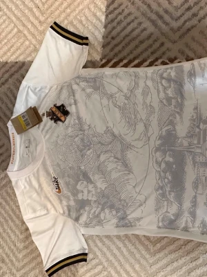 Vit AIK matchtröja Nike - Snygg vit matchtröja från Nike med AIK:s klubbmärke i svart och guld på bröstet. Tröjan har ett detaljerat grått mönster över hela framsidan och svarta/guldiga ränder på ärmsluten. Perfekt för dig som vill stötta AIK på match eller träning.