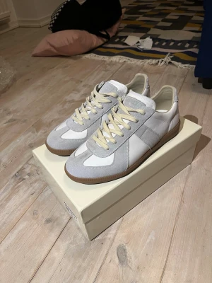 Maison Margiela Gats - Snygga sneakers från Maison Margiela i vitt och grått läder och mocka, med beigea snören och klassisk gummisula. Perfekt för dig som vill ha en clean och stilren look med premiumkänsla. Ikonisk design som passar till många. Skriv gärna frågor om det är något du undrar!