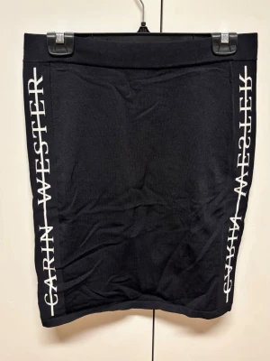 Svart kjol från Carin Wester - Svart kjol från Carin Wester med vita logotexter längs båda sidorna. Kjolen har en rak passform och bred resår i midjan. Perfekt för en trendig och sportig look.