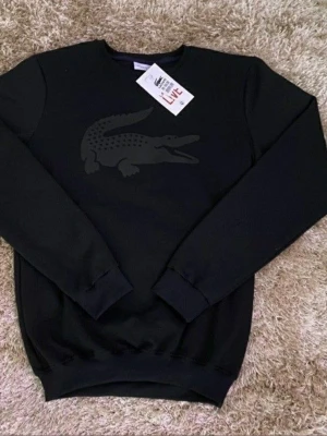 Svart sweatshirt från Lacoste - Cool svart sweatshirt från Lacoste med den klassiska krokodilloggan i svart på bröstet. Tröjan har rund halsringning, långa ärmar och ribbade muddar. Perfekt för en avslappnad och stilren look.