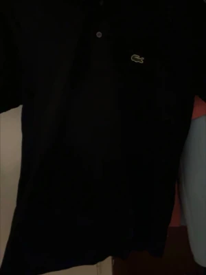 Svart pikétröja från Lacoste - Klassisk svart pikétröja från Lacoste med den ikoniska gröna krokodilloggan på bröstet. Tröjan har korta ärmar, krage och knappar framtill. Perfekt för en clean och stilren look.