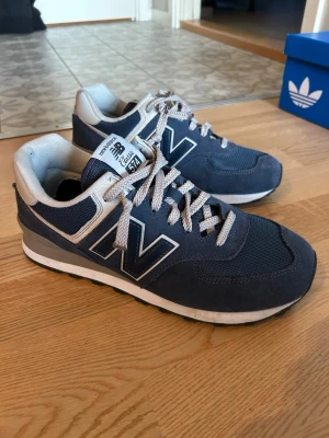 Blå New Balance 574 sneakers - New Balance sneakers, använda en gång. Försökte att justera skosnörena men skoformen passar ej min fot. Skosnörena är avklippta och brända i ändarna. Annars är skorna i nyskick.