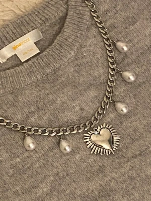 Kedjehalsband med pärlor och hjärta - Snyggt silverfärgat kedjehalsband rostfritt stål med droppformade pärlor och ett stort hjärthänge med solstråle-design. Perfekt statement-piece för att lyfta din outfit och ge en cool touch. Metallkedja med pärldetaljer och unikt hjärthänge.