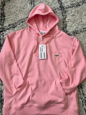 Rosa hoodie från Lacoste - Snygg rosa hoodie från Lacoste med klassisk krokodillogga på bröstet. Regular fit-modell med huva och dragsko samt stor magficka framtill. Tillverkad i mjuk bomullsblandning som känns skön mot huden. Perfekt för en avslappnad och trendig look.