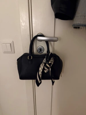 Svart handväska med scarfdetalj - Stilren svart handväska i boxig modell med två handtag och guldfärgade metalldetaljer. Väskan har en avtagbar svartvit scarf knuten runt handtaget och dragkedja upptill. Perfekt accessoar för att lyfta din outfit.
