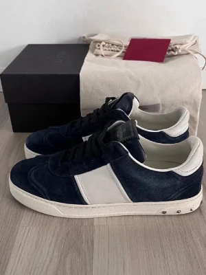 Valentino blåa och vita sneakers - Snygga sneakers från Valentino med mörkblå mocka och vita detaljer. Skorna har klassisk låg profil, svarta skosnören och en vit sula med metallnitar på hälen. Perfekt för dig som vill ha en exklusiv och stilren look.