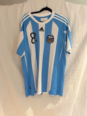 Argentina  2010 Verón #8 Adidas - Säljer en klassisk Argentina matchtröja från Adidas med ljusblå och vita ränder, nummer 8 och Verón på ryggen. Tröjan har officiellt AFA-märke, broderad Adidas-logga och Climacool-material som andas. Perfekt för fotbollsfans och samlare.