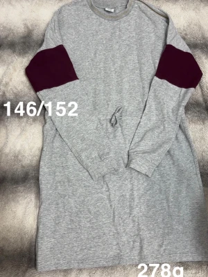 Grå sweatshirtklänning med vinröda detaljer - Mysig grå sweatshirtklänning med vinröda partier på ärmarna och knyt i midjan. Perfekt för en avslappnad stil.🌸 Använd en gång. Storlek 146/152