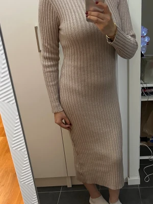 Beige ribbad stickad långklänning - Snygg beige stickad långklänning med ribbat mönster och lång ärm. Klänningen har en kroppsnära passform och hög hals, perfekt för en trendig och bekväm look.