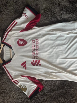 Liverpool bortatröja Ekitike 22 Adidas - Snygg Liverpool FC bortatröja från Adidas i vitt med vinröda detaljer och svarta inslag. Tröjan har tryck med Ekitike och nummer 22 på ryggen, samt Premier League-märke på ärmen. Materialet är väldigt lätt. 100% äkta, köpt på Liverpools hemsida och kvitto finns. (Aldrig använt)