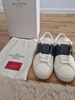 Valentino Garavani vita sneakers - Snygga vita sneakers från Valentino Garavani med bred mörkblå rem över mitten. Skorna har klassisk låg profil, vita snören och diskret logga på plösen. Tillverkade i läder med stilren design som passar till många outfits.
