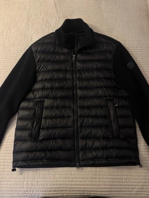 Svart hybridjacka från Colmar - Snygg svart pufferjacka från Colmar med stickade ärmar och vadderad framsida. Jackan har två dragkedjefickor och hög krage. Perfekt för kalla dagar och har en modern, sportig look med Colmar-logga på ärmen.