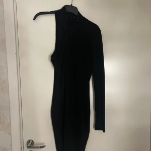 Svart oneshoulder kortklänning - Snygg svart kortklänning med oneshoulder-design och lång ärm. Klänningen har en tight passform och är tillverkad i ett stretchigt material som sitter bekvämt mot kroppen. Perfekt för dig som vill sticka ut med en unik och trendig look.