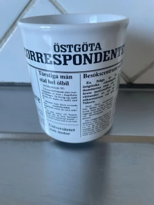 Mugg med tidningsmotiv Östgöta - Vit mugg i keramik med tryck av tidningssidor från Östgöta Correspondenten. Texten är svart och täcker större delen av muggen. Tillverkad i England. Perfekt för dig som gillar unika och roliga muggar med retrostil.