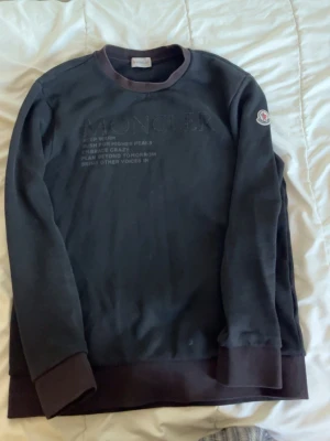 Svart sweatshirt från Moncler - Snygg svart sweatshirt från Moncler med mörkare muddar vid ärmslut, hals och nederkant. Diskret Moncler-logga på vänster ärm och broderad text på bröstet. Perfekt för dig som gillar stilrena och exklusiva plagg.