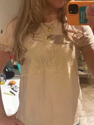 Beige blus med spetsdetaljer - Söt beige blus med korta ärmar och fina spetsdetaljer över bröstet och ärmsluten. Blusen har en mjuk och feminin look, perfekt att styla med jeans eller kjol. Materialet känns lätt och luftigt.