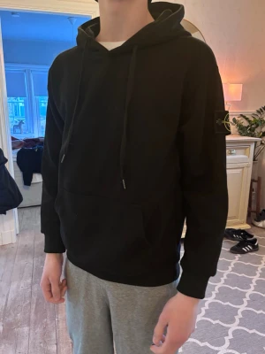 Svart hoodie från Stone Island stl S - Säljer en svart hoodie från Stone Island med klassisk patch på vänster ärm. Hoodien har huva med dragsko, magficka och ribbade muddar. Tillverkad i mjuk bomull för skön känsla och avslappnad stil. Det är storlek S på tröjan och killen på bilden är 180 cm 62 kg. Tröjan är använd fåtal gånger så mycket gott skick!