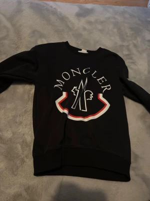 Svart Moncler sweatshirt med tryck - Snygg svart sweatshirt från Moncler med stort vitt och rött logotryck på bröstet. Tröjan har rund halsringning och långa ärmar. Perfekt för dig som gillar streetwear och vill sticka ut med ett exklusivt märke.
