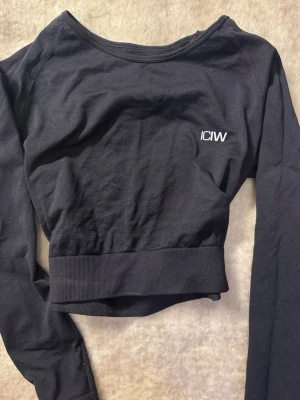 Svart cropped tröja från ICANIWILL - Snygg svart cropped långärmad tröja från ICANIWILL med vit logga på bröstet. Tröjan har bred ribbad mudd i midjan och är tillverkad i mjukt, stretchigt material som passar perfekt till träning eller chill.