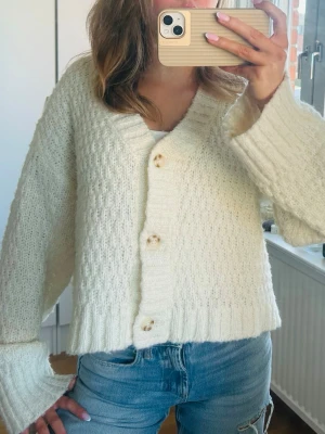 Vit stickad kofta med stora knappar - Mysig vit stickad kofta med oversized passform och stora dekorativa knappar framtill. Koftan har breda ribbade muddar och ett diskret mönster i stickningen. Perfekt att slänga på sig över en t-shirt och jeans.