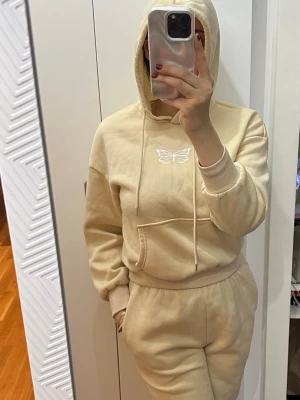 Beige mjukisdress med fjärilstryck - Beige loungewear-set med hoodie och matchande byxor. Hoodien har dragsko, magficka och ett vitt fjärilstryck på bröstet. Byxorna har resår i midjan och är gjorda i mjukt material, perfekt för chill dagar hemma.