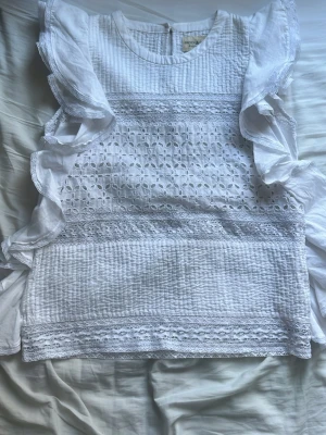 Vit blus med volanger och spets - Supersöt vit blus från Ralph Lauren med broderade spetsdetaljer och volanger längs ärmhålen. Blusen har rund halsringning och är tillverkad i ett luftigt bomullstyg med broderade blommor och struktur. Perfekt för en fräsch och somrig look.