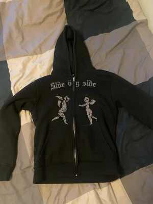 Rhinestone hoodie - Säljer en svart hoodie med dragkedja och huva. På bröstet finns två änglar och texten 'side by side' i rhinestone som ger ett unikt och edgy utseende. Alla stenar ser ut att sitta kvar möjligen någon emstaka som kan vara av men inget märkbart! Storlek xxs sitter mer som xs. 
