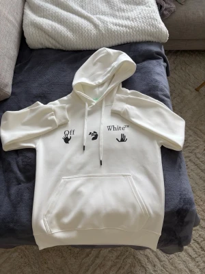 Vit Off-White hoodie med blått tryck - Säljer en vit hoodie från Off-White med svart tryck och handmotiv på bröstet samt ett stort blått grafiskt tryck på ryggen. Hoodien har huva med dragsko och en stor magficka. Perfekt för dig som gillar streetwear och vill sticka ut. Priset kan diskuteras!