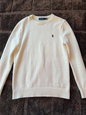 Vit stickad tröja från Polo Ralph Lauren - Klassisk vit stickad tröja från Polo Ralph Lauren med rund halsringning och diskret mörkblå logga broderad på bröstet. Tröjan har ribbade muddar vid ärmslut och nederkant. Perfekt för en clean och stilren look.