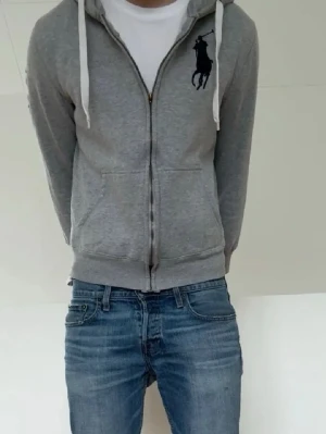 Grå zip hoodie från Ralph Lauren big pony - Snygg grå hoodie från Ralph Lauren med dragkedja och stor svart logga på bröstet. Hoodien har vita snören i huvan och två fickor framtill. Perfekt för en chill och avslappnad stil. Materialet känns mjukt och bekvämt. storlek är runt xs-s