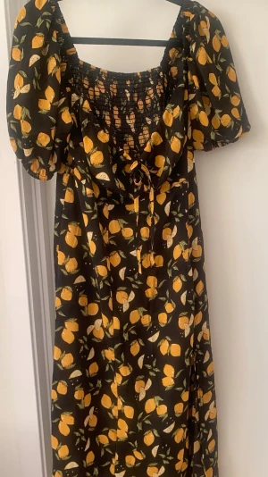 Svart klänning med citronmönster - Söt svart klänning med gula citroner och gröna blad, puffärmar och smockad överdel. Klänningen har knytning framtill och en luftig, flowy passform. Perfekt för dig som gillar färgglada och lekfulla prints. Oanvänd och i väldigt bra skick 