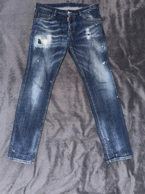  Äkta Dsquared2 blå slitna jeans - Snygga blå jeans från Dsquared2 med slitningar och distressed detaljer framtill. Jeansen har smal passform och klassisk femficksdesign. Orange Dsquared2-logga syns på baksidan i linningen. Perfekta för en trendig streetstyle-look. Passar M