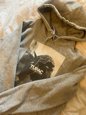 Grå hoodie med Tupac-tryck - Snygg grå hoodie med stort svartvitt Tupac-tryck på bröstet. Klassisk modell med huva och dragsko samt känguruficka framtill. Perfekt för dig som gillar streetstyle och vill ha en bekväm och stilren tröja med ikoniskt motiv.