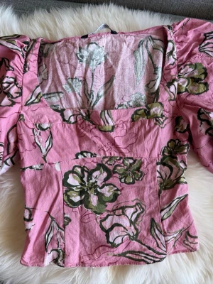 Rosa blommig blus med puffärm - Supersöt rosa blus med grönt och vitt blommönster. Blusen har puffiga korta ärmar och fyrkantig ringning. Perfekt för dig som vill ha en feminin och trendig topp till sommaren. Materialet känns lätt och luftigt.